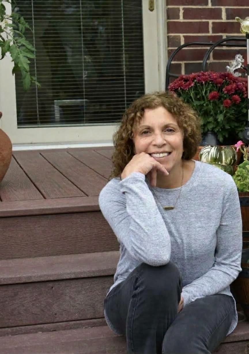 Smiling woman with curly brown hair resting her chin on her hand, sitting on outdoor wooden deck stairs with potted plants and brick wall in the background.