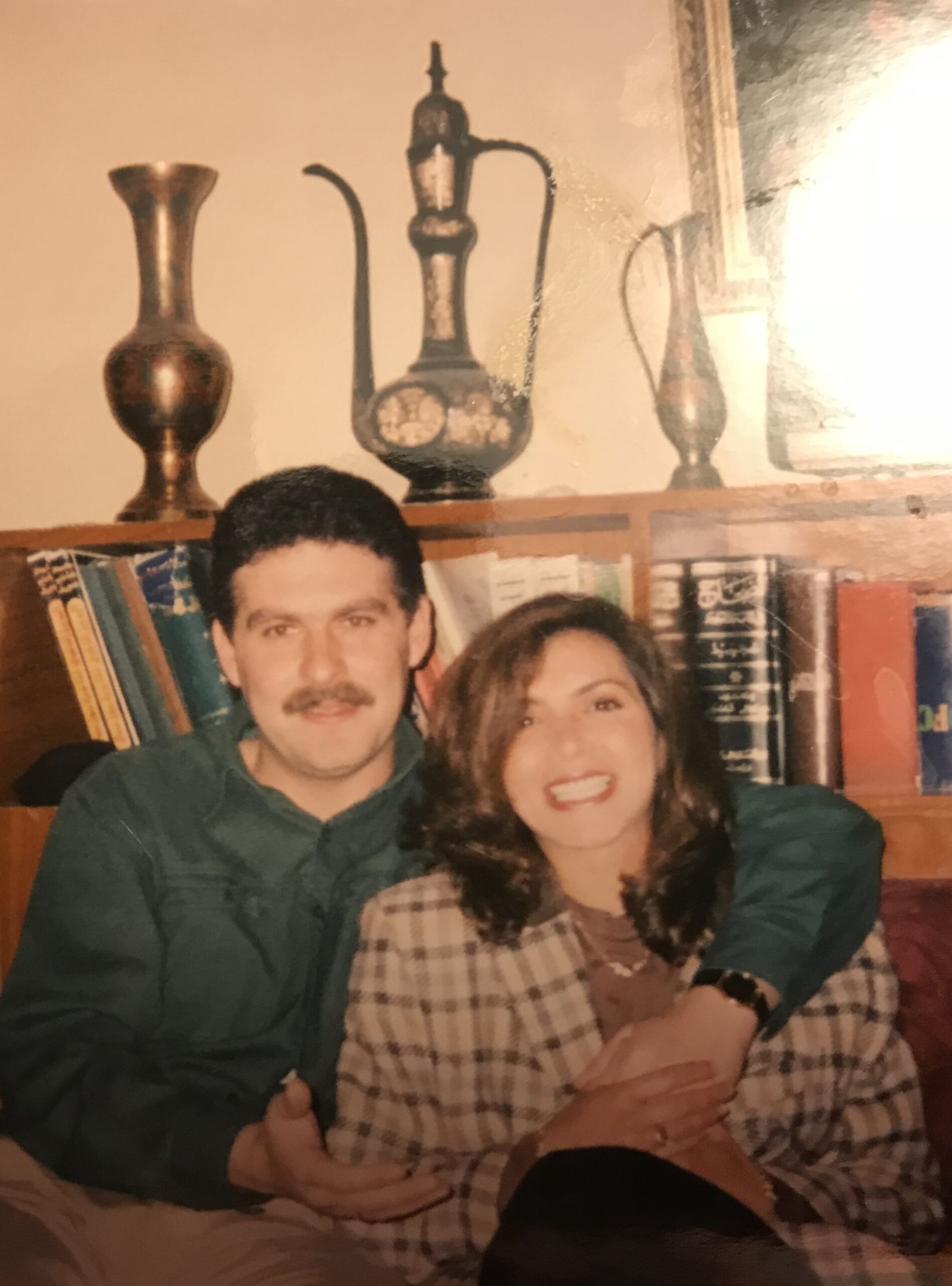 a man with short dark hair and a mustache wearing a dark green button-down shirt, and a woman with voluminous wavy brunette hair and a bright smile wearing a cream and brown plaid blazer, sitting closely together with his arm around her, in a warm indoor setting with a wooden bookshelf and ornate brass Middle Eastern pitchers in the background, vintage photograph aesthetic